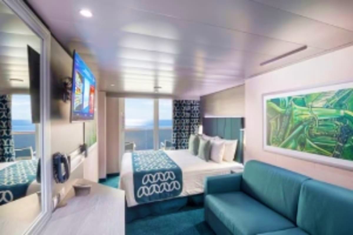 Modern cruise ship cabin with ocean view, bed, teal sofa, and floor-to-ceiling windows overlooking the sea