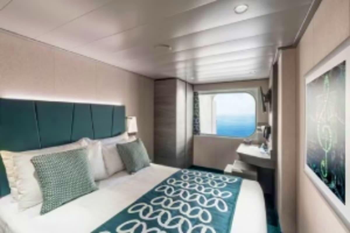 Modern cruise ship cabin interior with ocean view window, queen bed with teal headboard, and contemporary furnishings