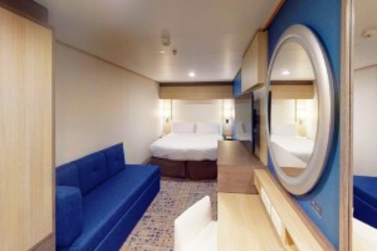 Modern cruise ship cabin interior with blue upholstered sofa, queen bed, round porthole window, and wood accents