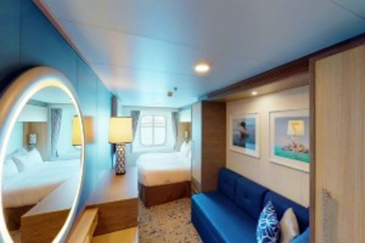 Modern cruise ship cabin with illuminated round mirror, queen bed, blue sofa, and ocean view porthole window