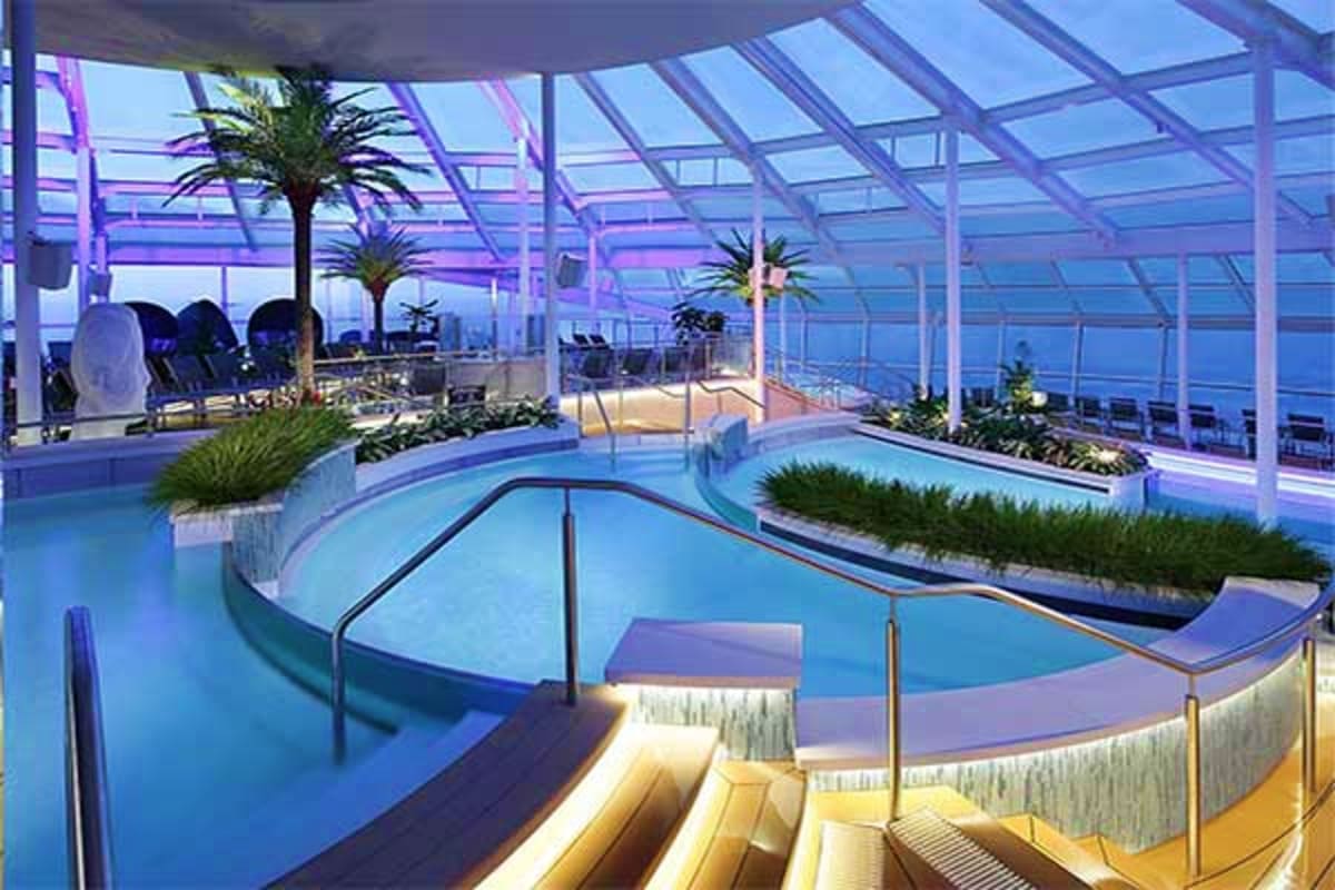 Modern cruise ship indoor pool area with curved lap pool, tropical plants, palm trees, and floor-to-ceiling windows overlooki