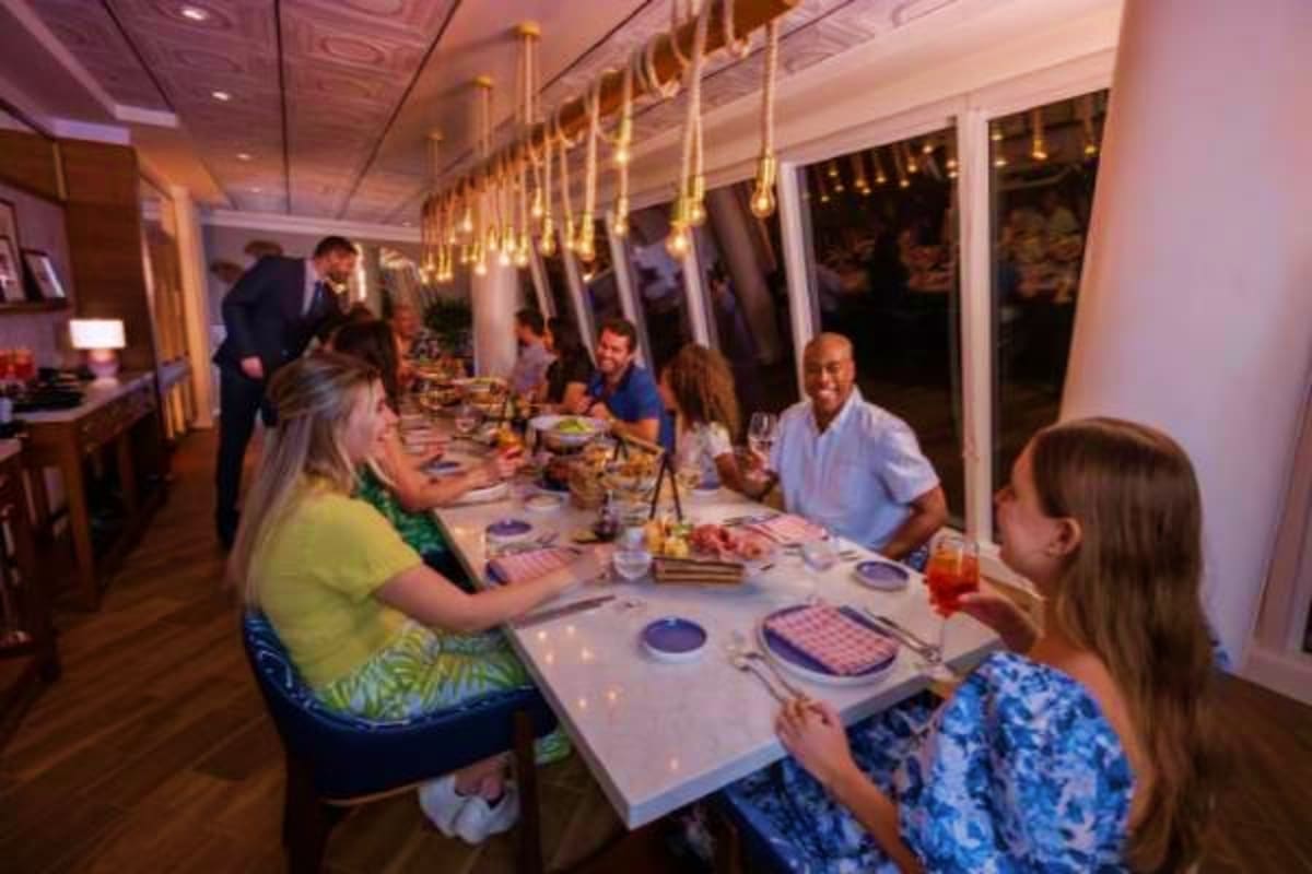 Passengers dining at long table in elegant cruise ship restaurant with gold pendant lights and ocean views through windows at