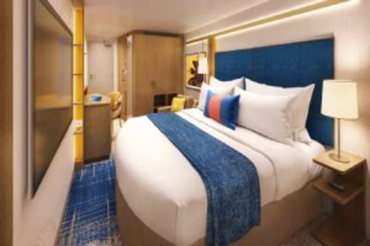 Modern cruise ship interior stateroom with queen bed, blue upholstered headboard, white bedding, decorative pillows, wooden w