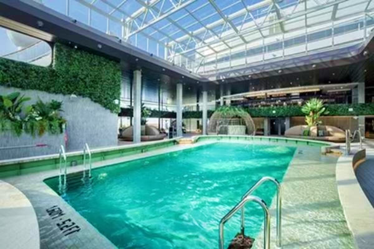 Indoor swimming pool with turquoise water, metal railings, glass ceiling, white columns, and tropical plants on cruise ship M