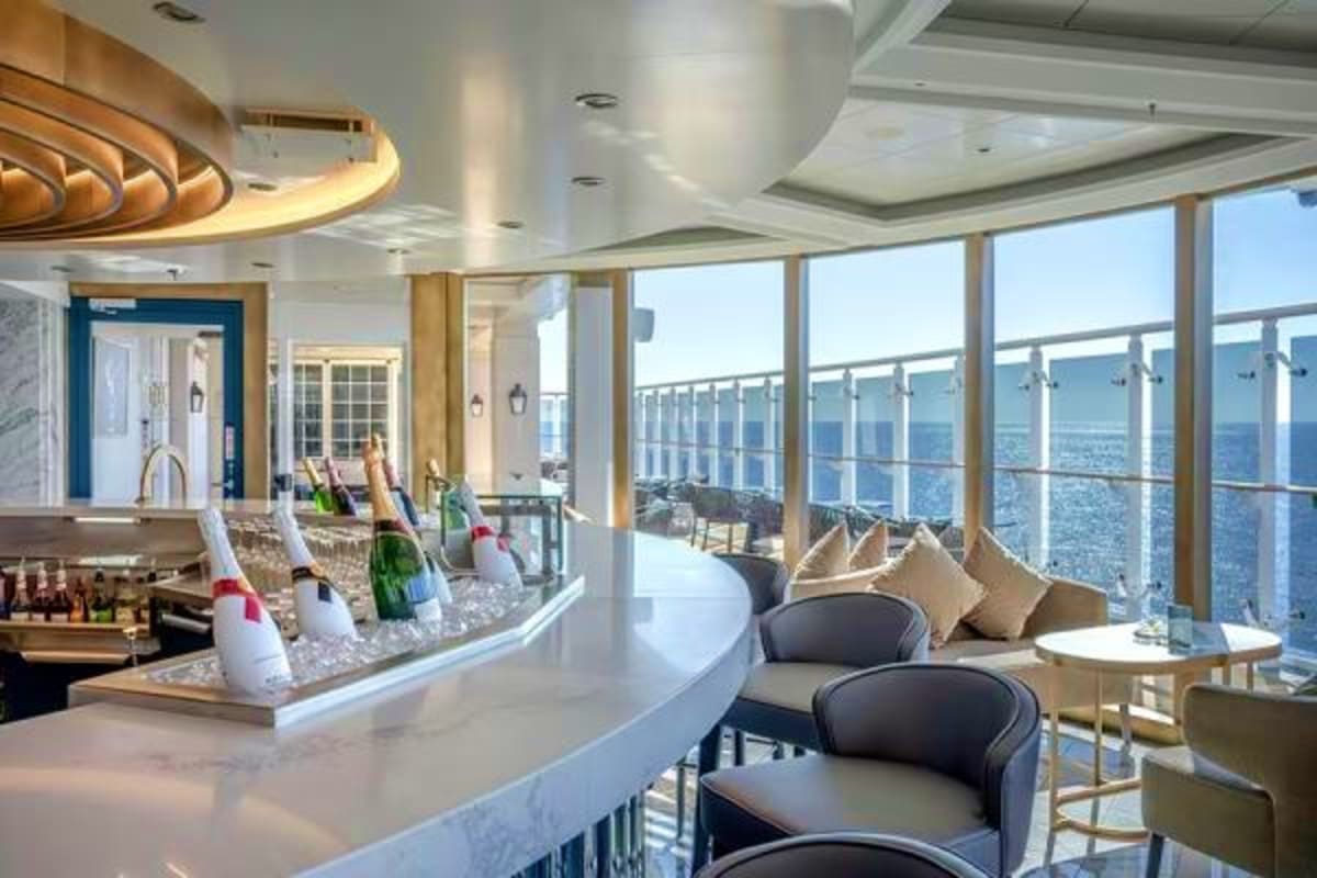Luxury cruise ship suite with curved bar, champagne bottles, panoramic ocean views through floor-to-ceiling windows, and mode