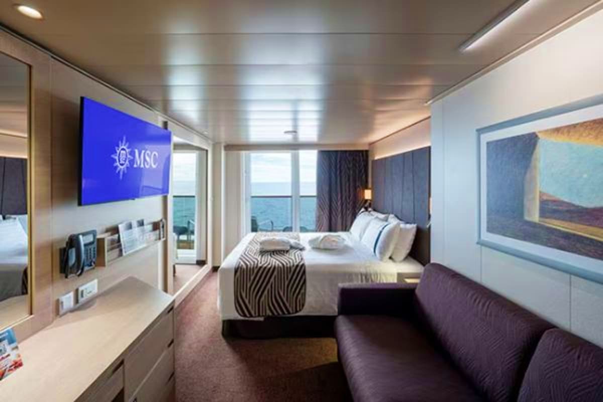Modern cruise ship cabin with queen bed, flat-screen TV showing MSC logo, ocean view balcony, and contemporary furnishings