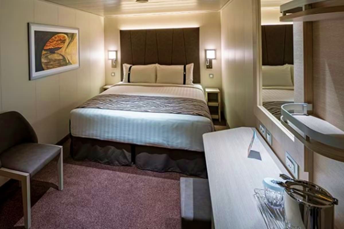 Modern cruise ship stateroom with queen bed, dark headboard, beige bedding, desk with amenities, and contemporary lighting