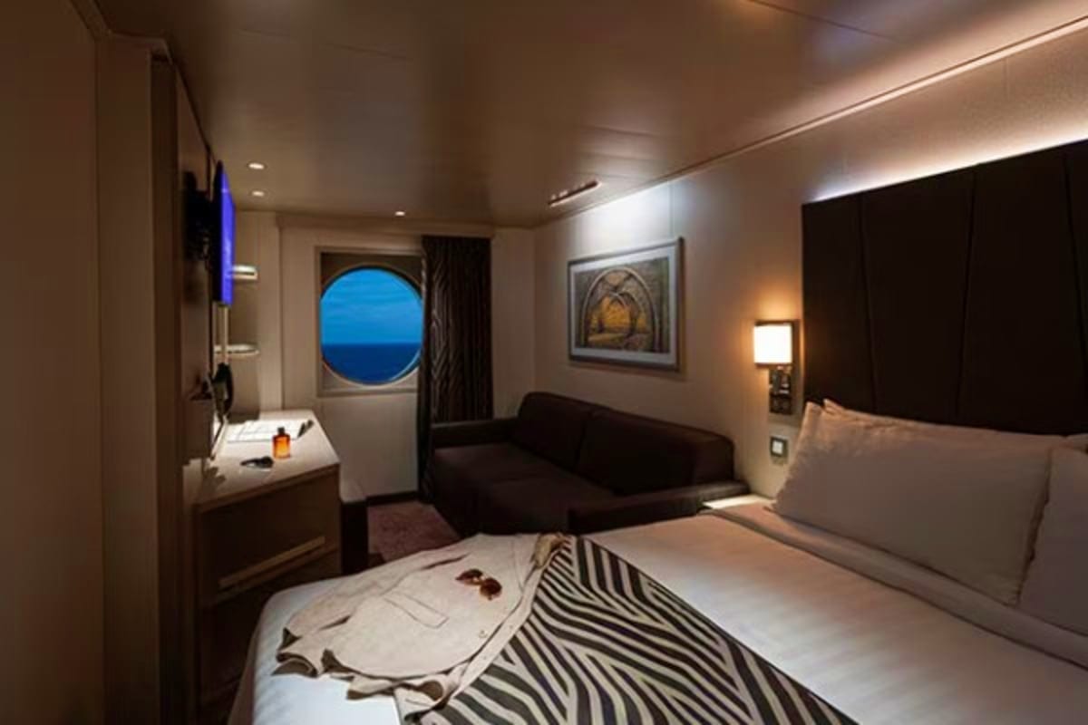 Luxury cruise ship cabin with porthole window showing ocean at dusk, queen bed with zebra print blanket, sofa seating, and mo