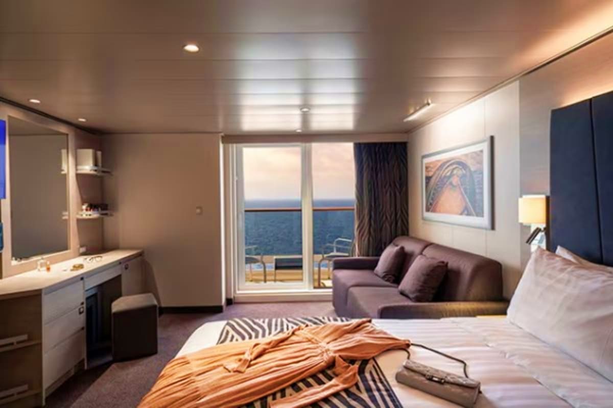 Luxury cruise ship cabin with ocean view balcony, bed, sofa seating area, and modern bathroom with vanity