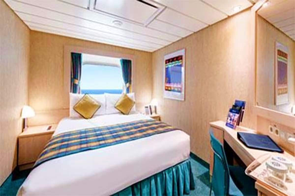 Modern cruise ship interior cabin with ocean view window, queen bed with gold pillows, desk workspace, and blue-green accents