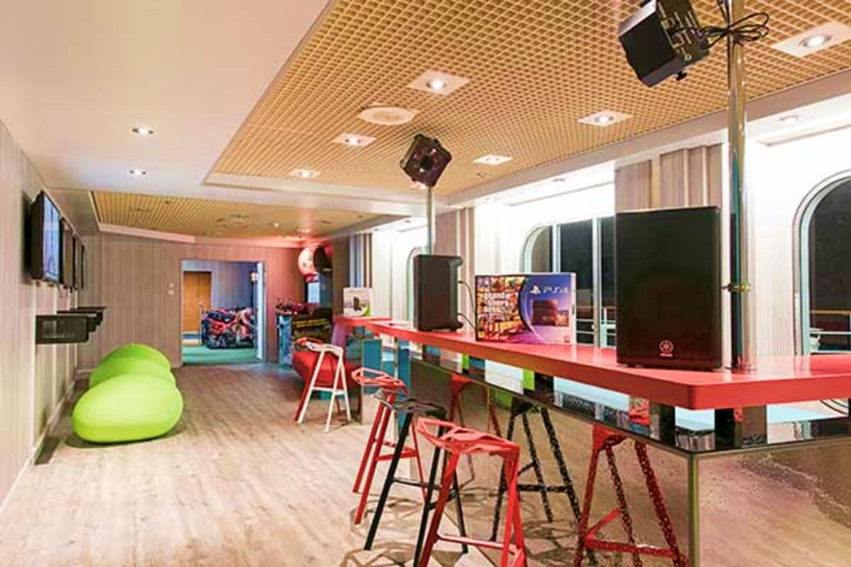 Modern cruise ship gaming lounge with red bar stools, red countertop, arcade games, green bean bag chair, and ceiling-mounted