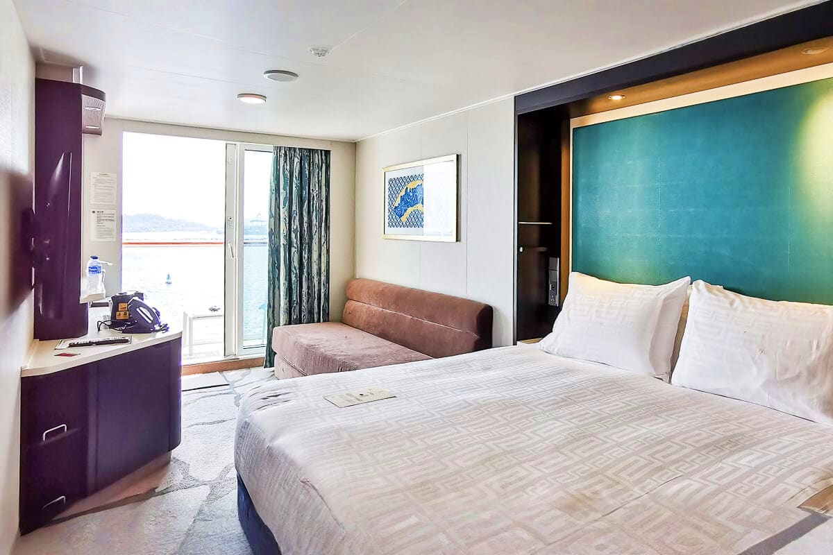 Modern cruise ship stateroom with queen bed, sofa, balcony ocean view, and teal accent wall headboard