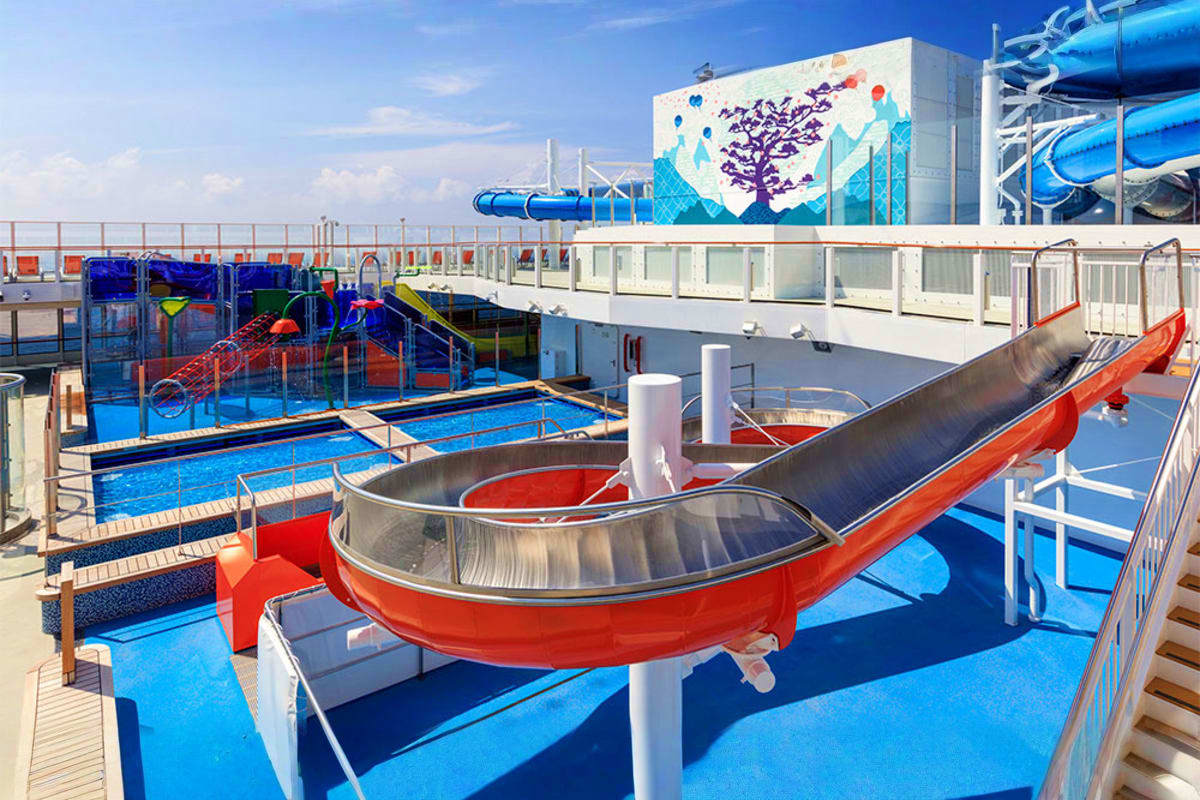 Cruise ship waterpark deck with red and black tube slides, blue pool areas, and colorful children's play structures under cle