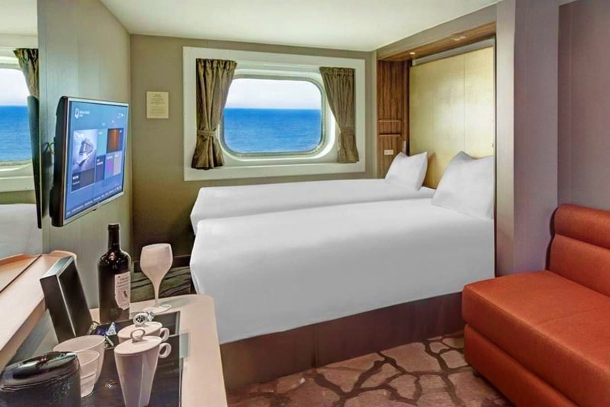Luxurious cruise ship cabin with twin beds, porthole ocean view, flat-screen TV, wine bottle, and modern furnishings