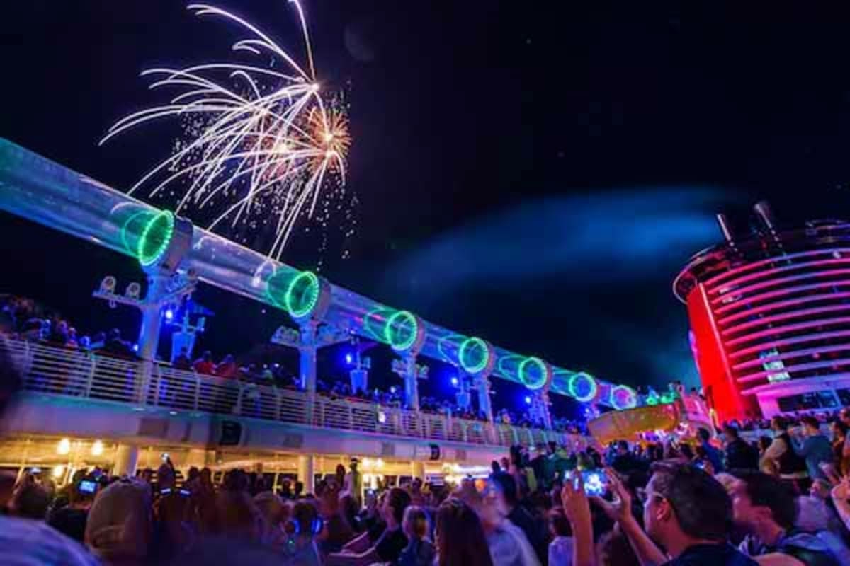 Fireworks explode above the Disney Dream cruise ship deck at night, with colorful green and blue lighting illuminating the sh