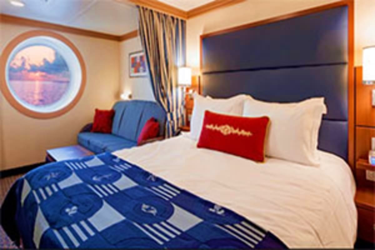 Luxury cruise ship stateroom with ocean view porthole, black headboard bed with red pillows, blue patterned bedding, and seat