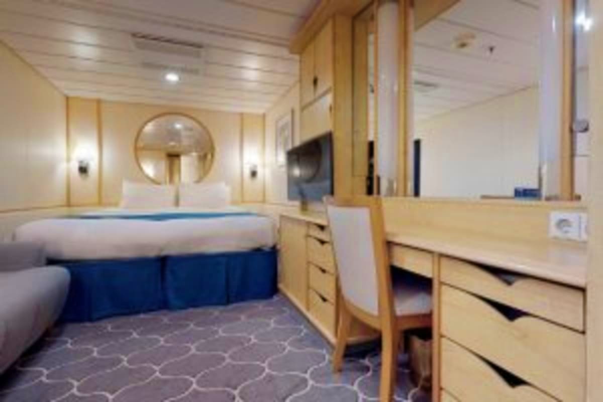 Modern cruise ship cabin with queen bed, wooden furnishings, vanity desk, circular mirror, and contemporary lighting