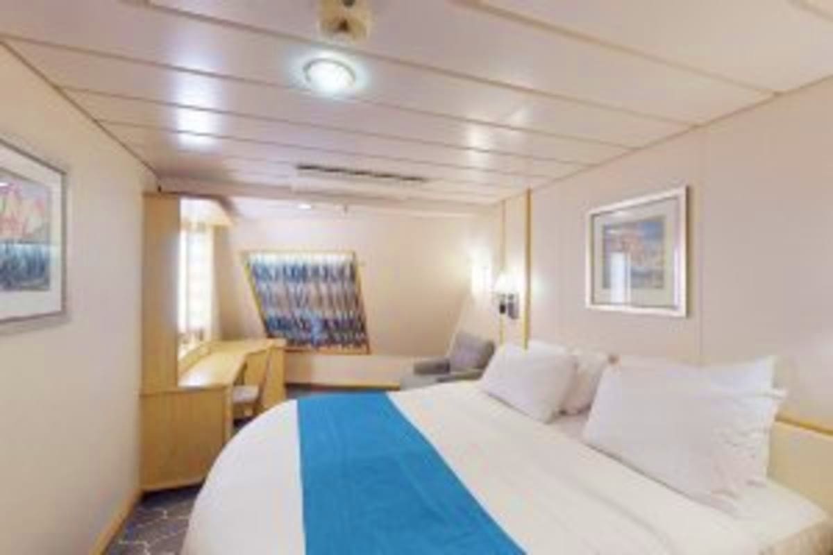 Modern cruise ship interior stateroom with queen bed, blue bed runner, white linens, wooden furniture, mirror, framed artwork