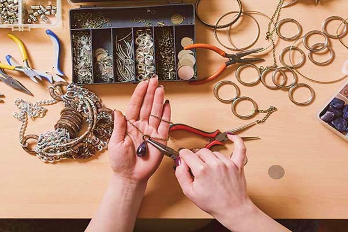 Jewelry maker's hands using pliers to create handmade accessories with beads, chains, and metal components on craft table