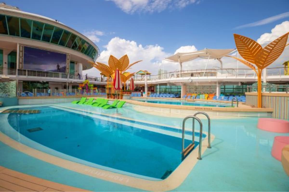Modern cruise ship aquatic center with Olympic-sized swimming pool, water slides, and lounge chairs under clear sky