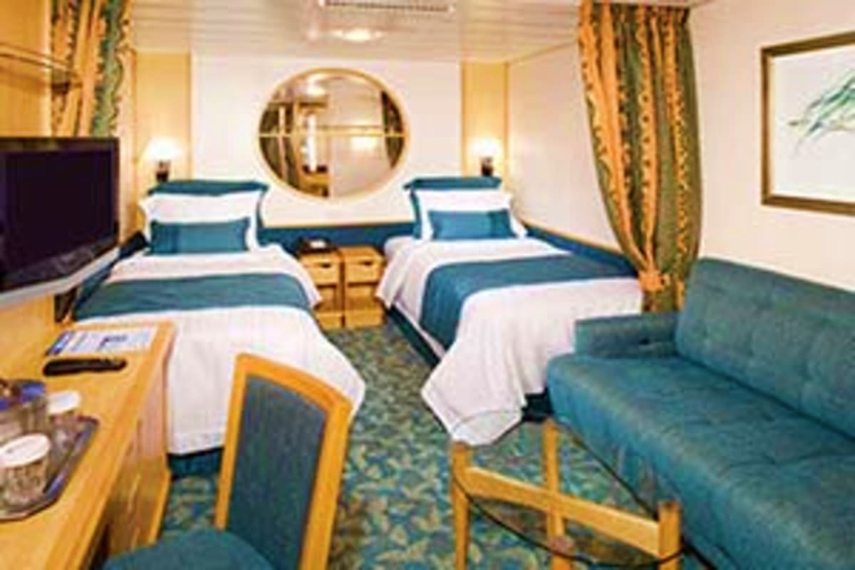Luxurious cruise ship cabin with two beds, teal and white bedding, wooden furniture, round mirror, sofa, and gold curtains