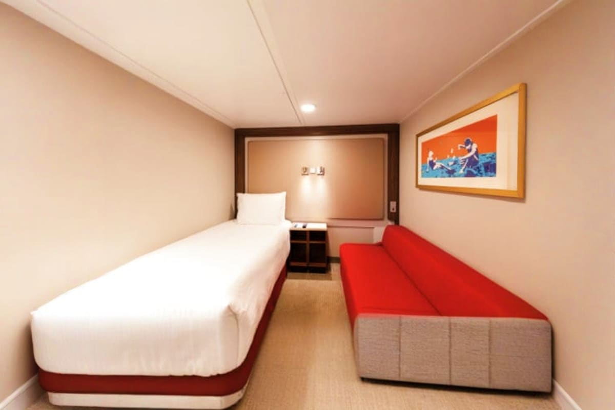 Compact cruise ship interior cabin with single bed, red sofa, nightstand, and framed artwork on beige walls