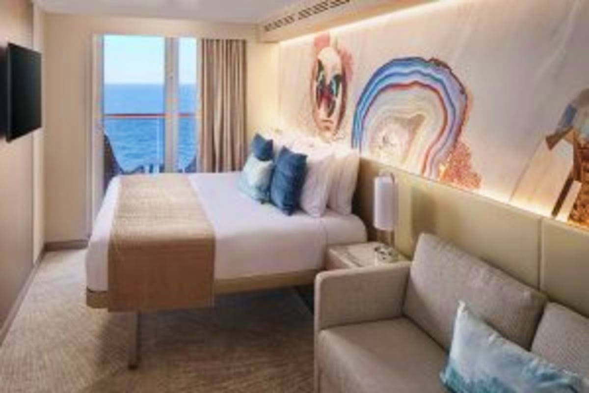 Modern cruise ship cabin with ocean view balcony, queen bed with blue pillows, sitting area, and abstract wall art overlookin