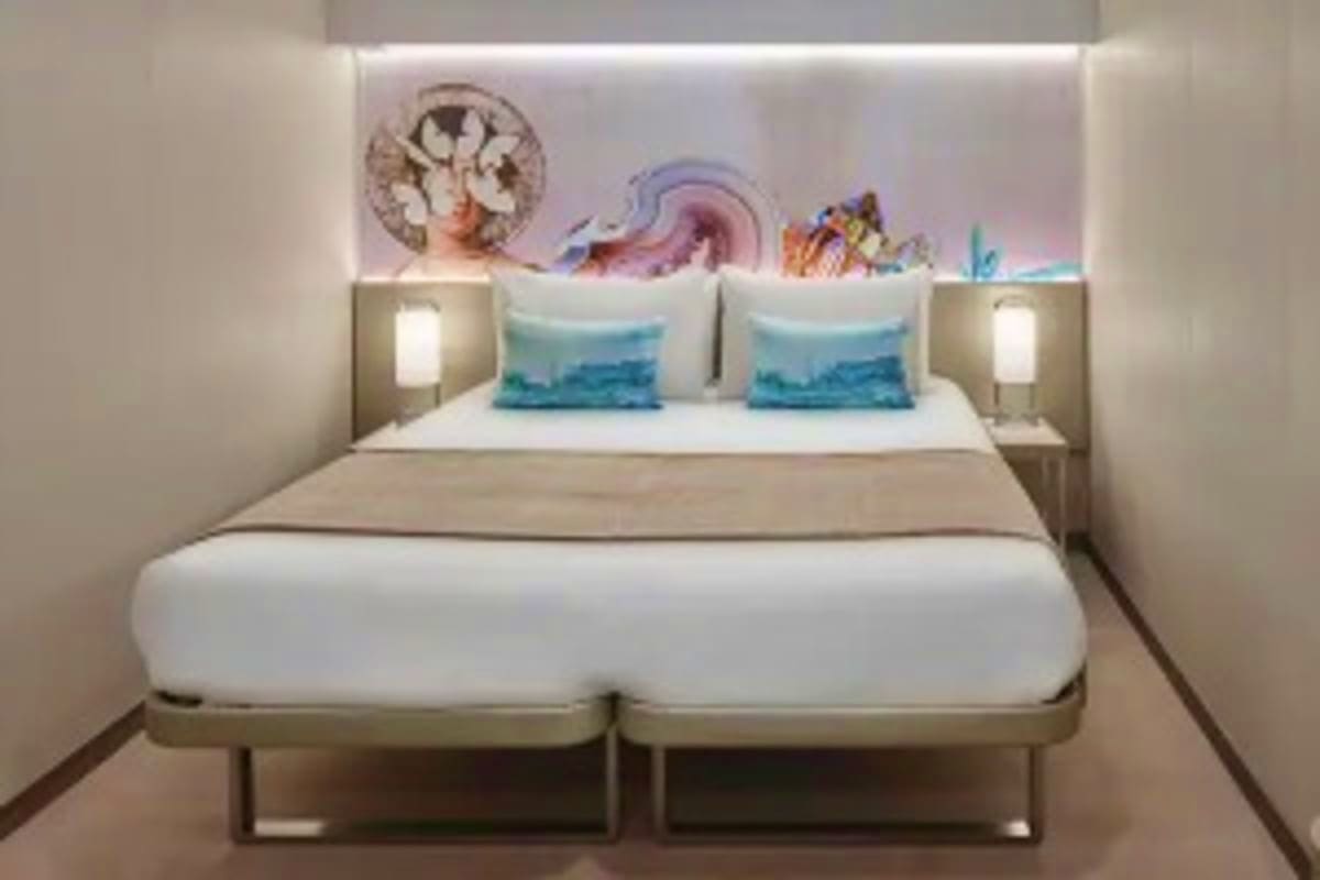 Modern cruise ship cabin with two twin beds, turquoise pillows, artwork of Greek islands and ocean scenes above headboard, am