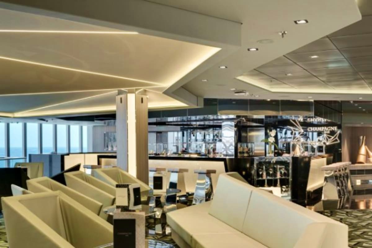 Modern cruise ship lounge with cream seating, ambient ceiling lighting, floor-to-ceiling windows, and contemporary black and 