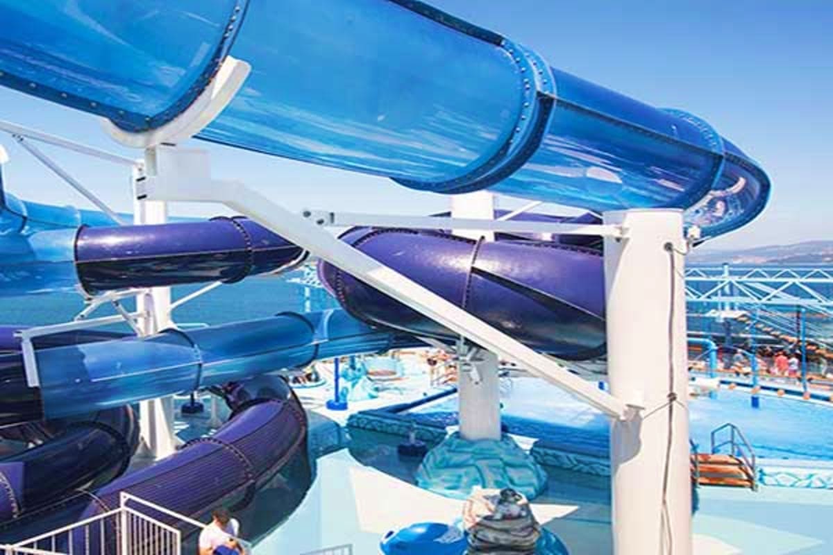 Blue water slides at a cruise ship pool deck with swimmers and leisure facilities visible below