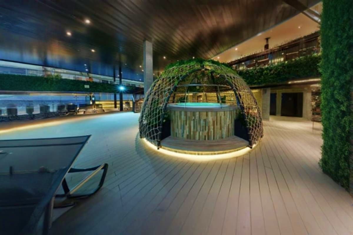 Modern cruise ship deck with illuminated woven pod structure, lounge chairs, and indoor pool area at night
