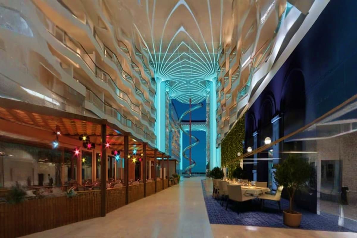 Modern cruise ship atrium with turquoise geometric ceiling design, restaurants with warm lighting, and ocean view through lar