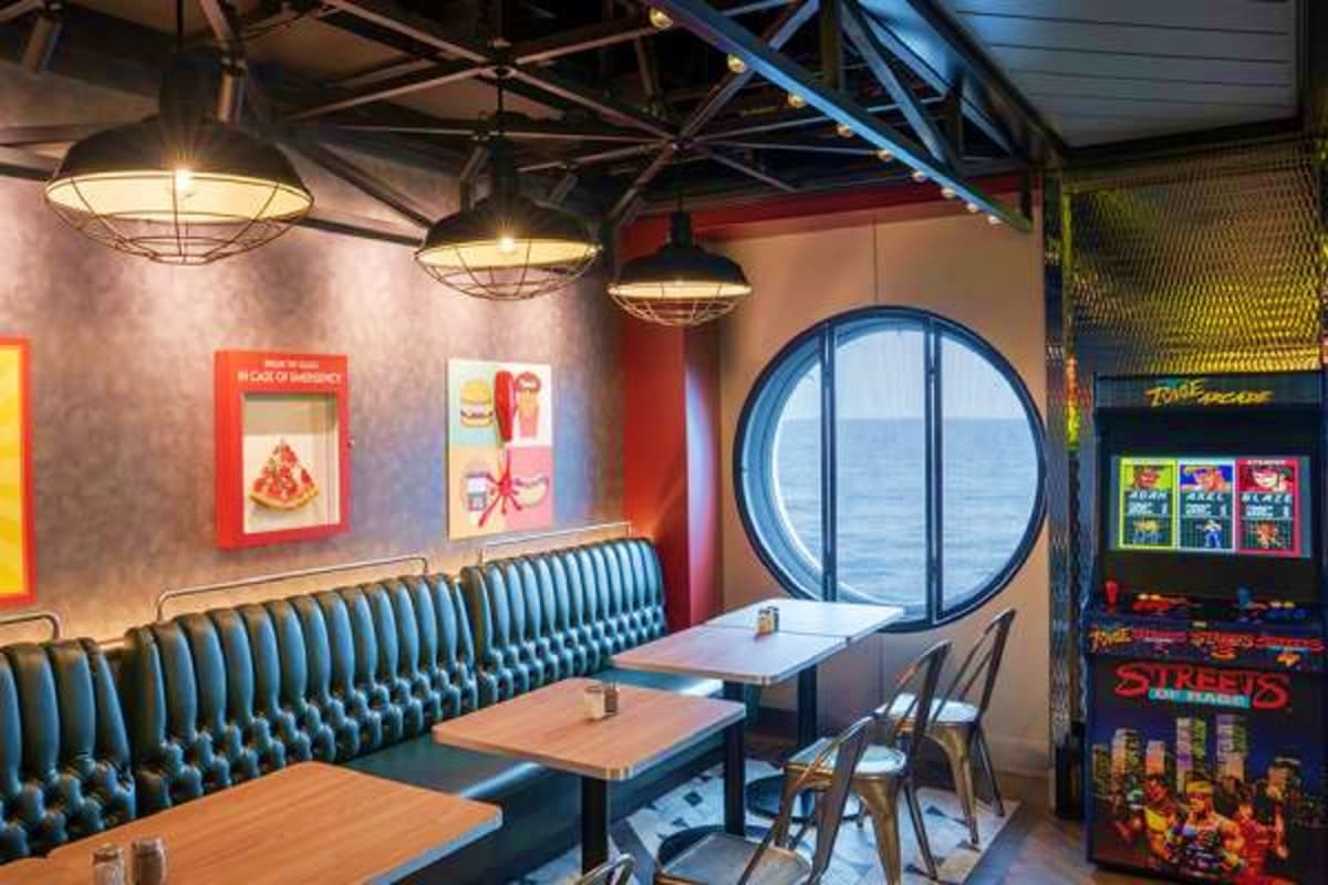 Modern retro-style restaurant interior with booth seating, pendant lighting, round porthole window, arcade game, and food pro