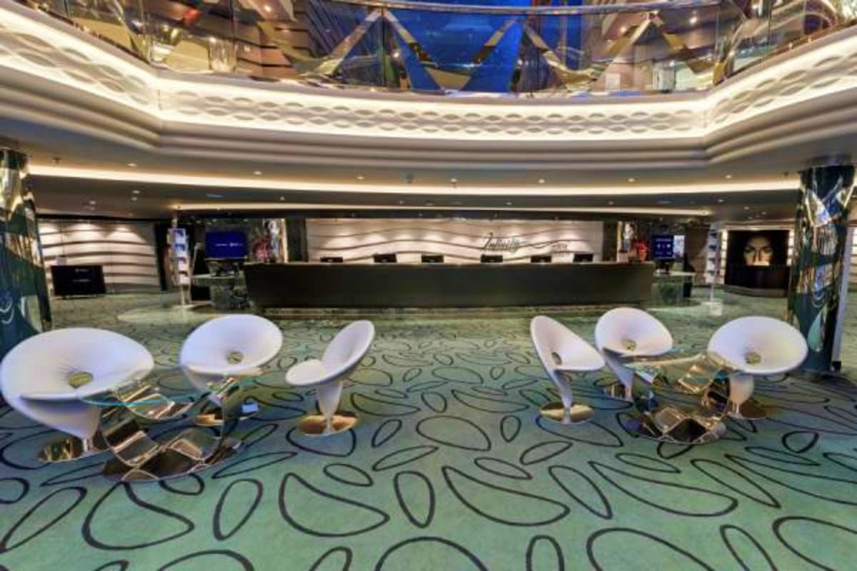 Modern cruise ship atrium with curved white pod seating, patterned carpet, and multi-level circular balconies overlooking the