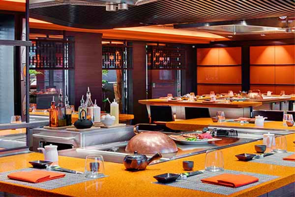 Modern cruise ship restaurant with orange and black color scheme, open kitchen with chef's counter, and elegant dining seatin