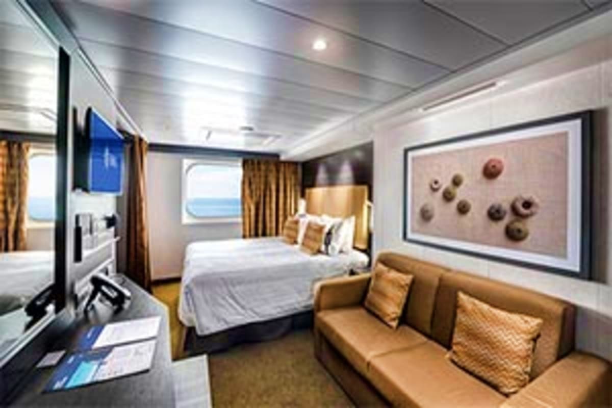 Luxury cruise ship stateroom with queen bed, seating area, flat-screen TV, and ocean view porthole window