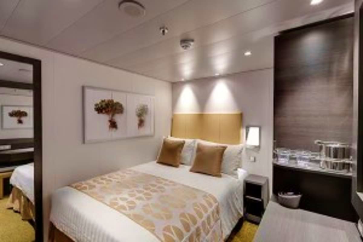 Modern cruise ship cabin with queen bed, gold accent headboard, botanical artwork, and ensuite bathroom with dark tile accent