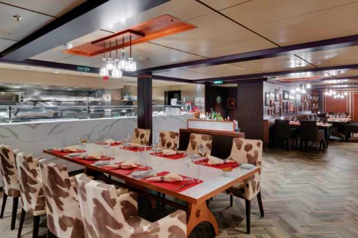 MSC Grandiosa Mediterranean Cruise Modern restaurant dining area on MSC Grandiosa with long wooden table, patterned chairs, red placemats, and open kitchen visi