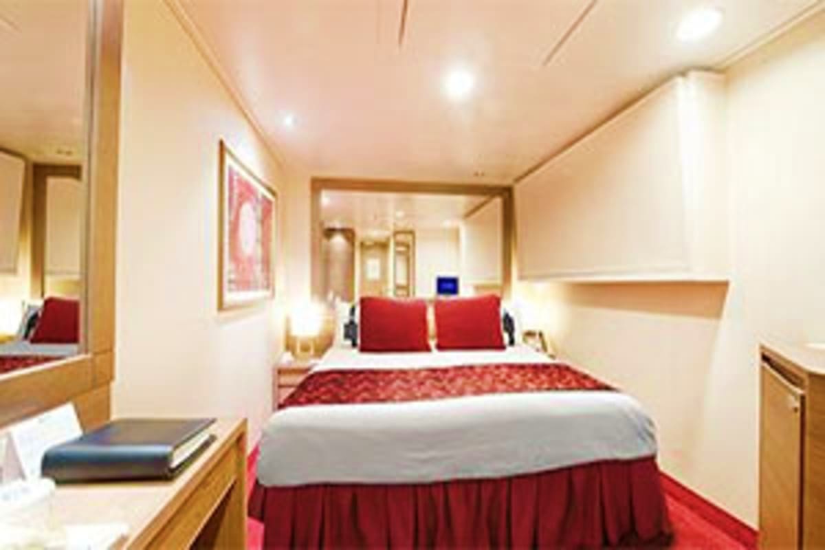 Modern cruise ship stateroom with queen bed, red bedding, wood furnishings, and mirrored vanity area