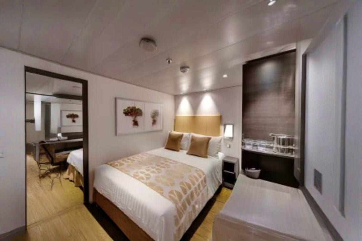 Modern cruise ship cabin with queen bed, gold accent headboard, white bedding, framed botanical artwork, ensuite bathroom wit