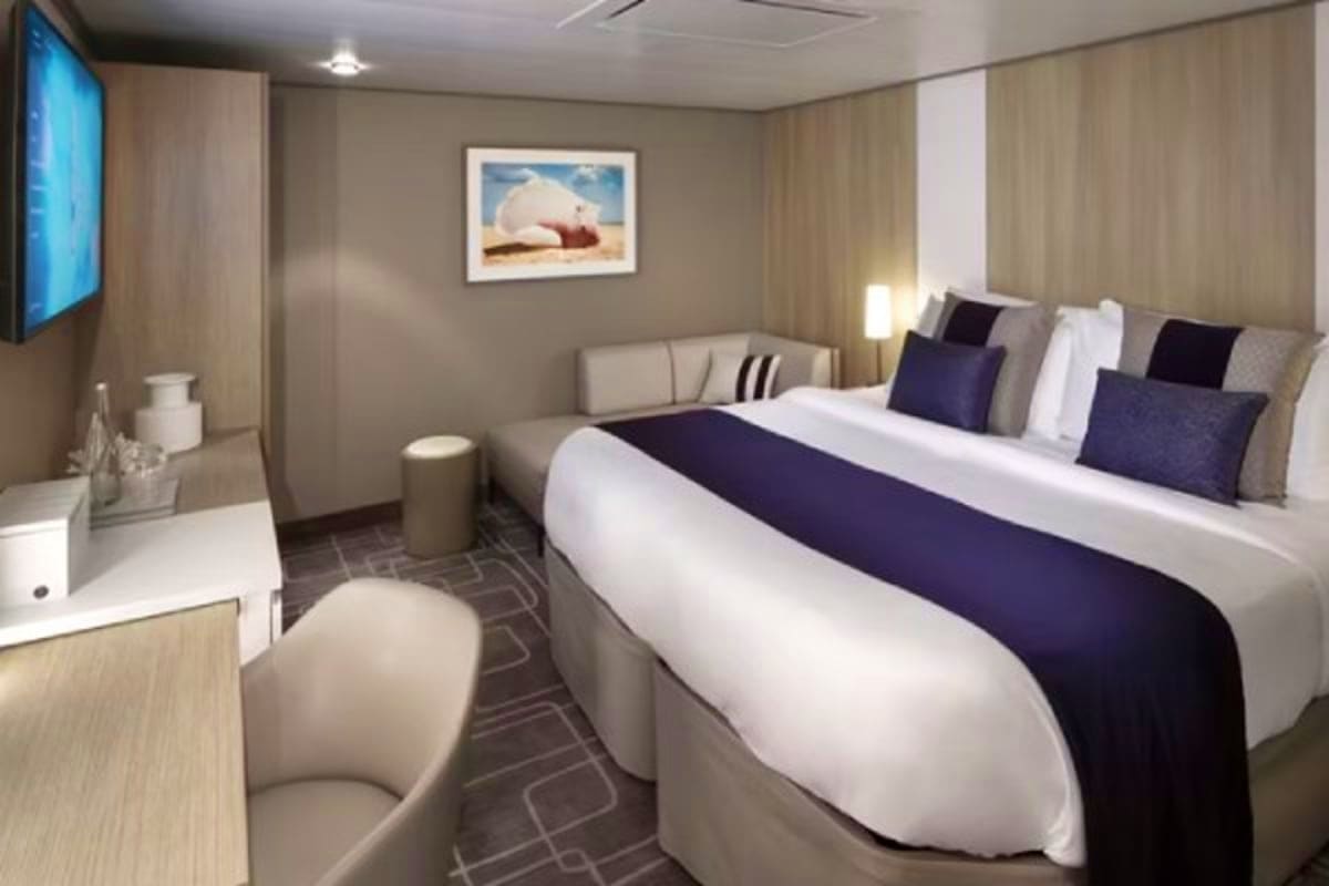 Modern cruise ship stateroom with queen bed, navy and white bedding, flat-screen TV, desk, and framed beach artwork on neutra