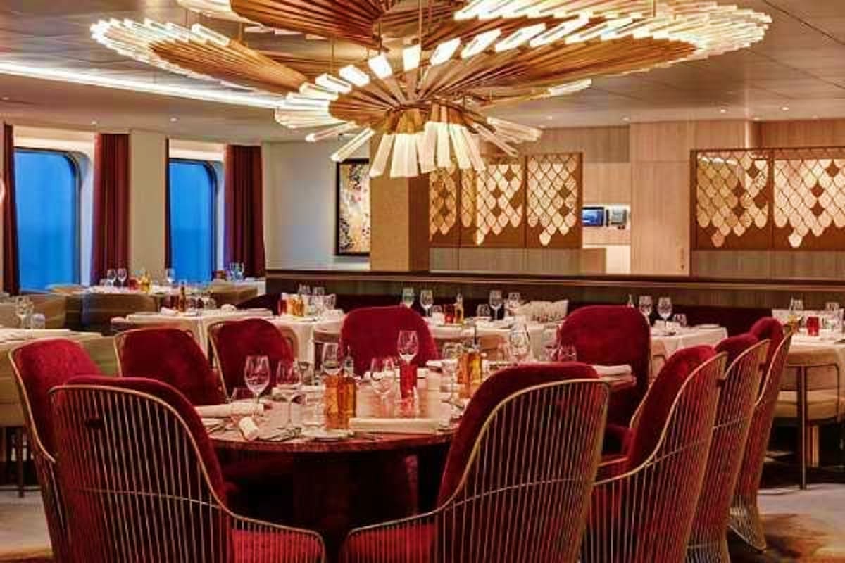 Elegant cruise ship dining room with red velvet chairs, round tables set for dinner, wooden ceiling fixture, and ocean view w