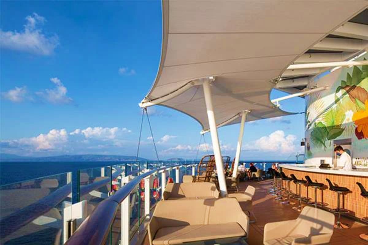 Outdoor deck on Celebrity Edge cruise ship with sun loungers, shade canopies, ocean views, and bar area overlooking blue wate