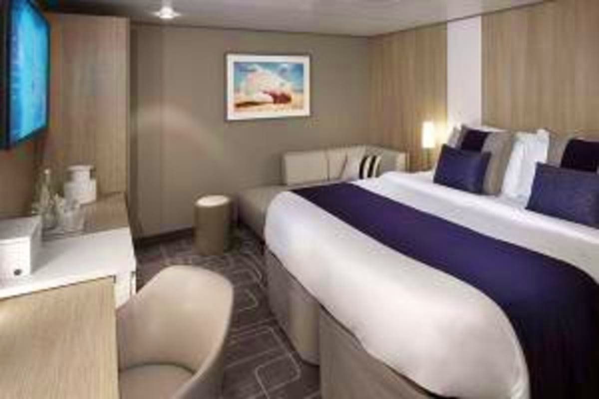 Modern cruise ship cabin with queen bed, sofa seating area, flat-screen TV, and neutral décor with ocean artwork