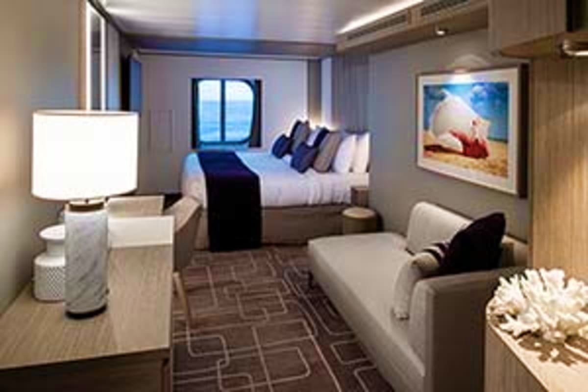 Modern cruise ship cabin with bed, sofa, porthole window, desk lamp, and framed beach artwork on wall