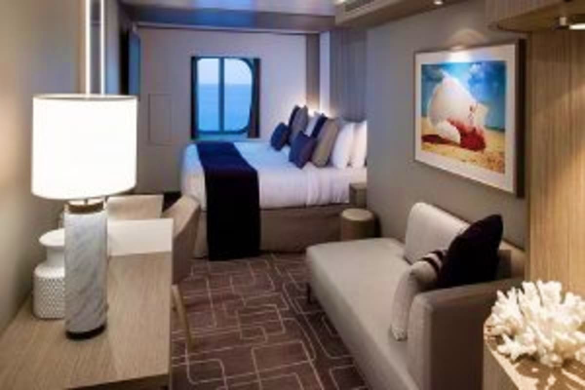 Modern cruise ship stateroom with queen bed, seating area, porthole window with ocean view, and contemporary furnishings