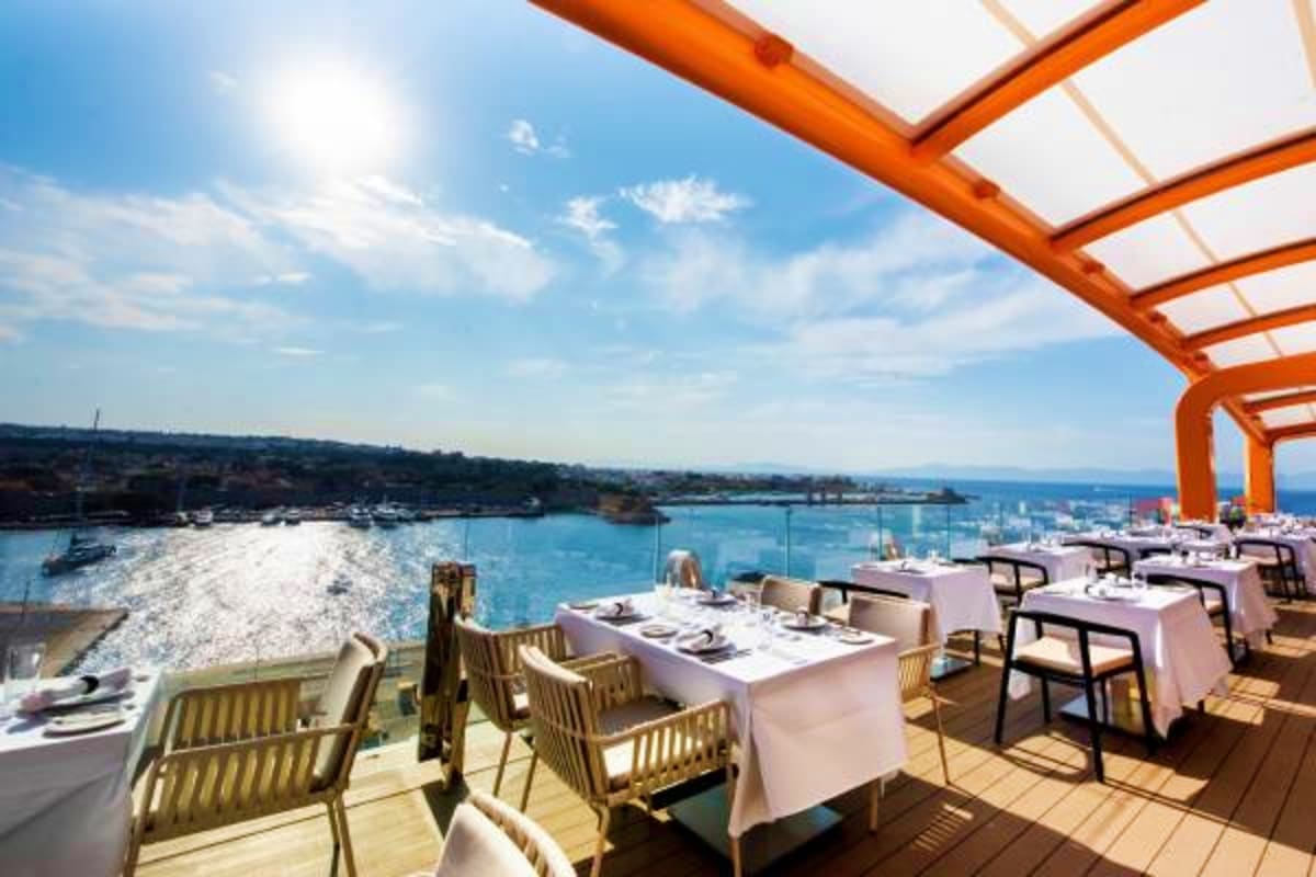 Oceanfront dining terrace on Celebrity Xcel cruise ship with orange pergola, white tablecloths, and Mediterranean coastal vie