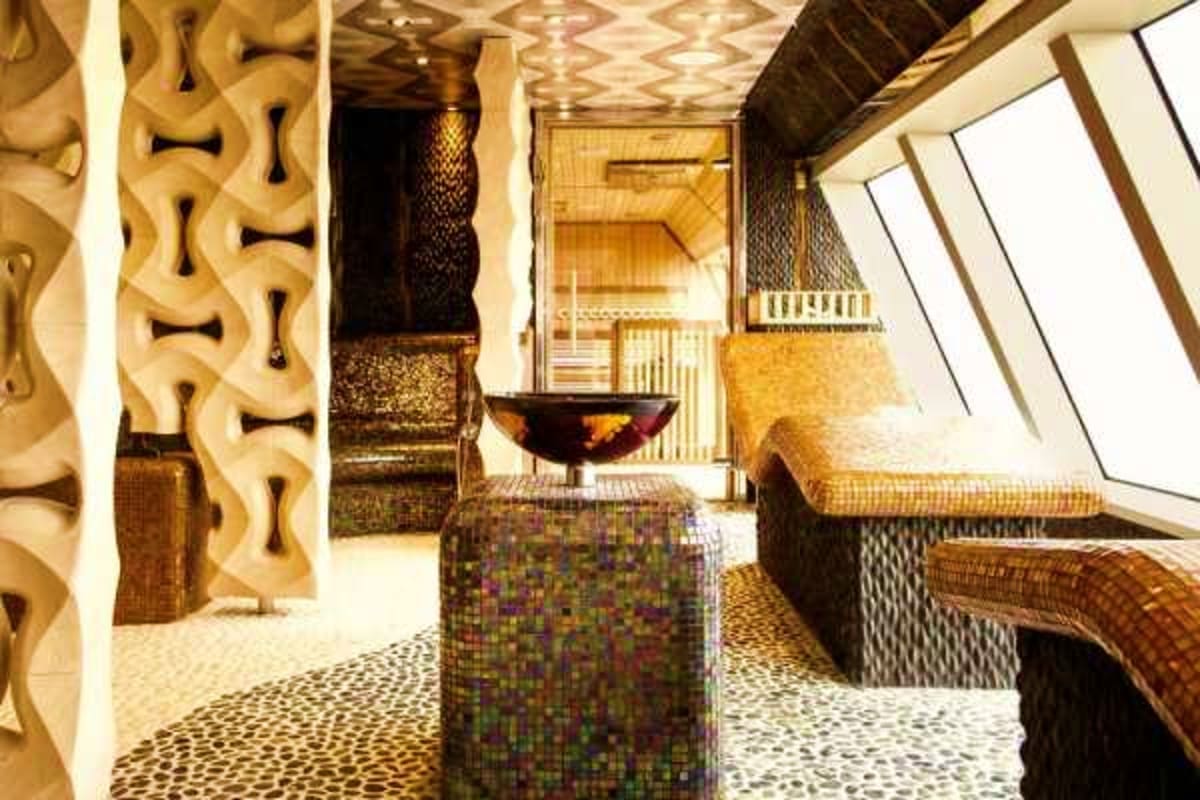 Luxury cruise ship spa interior with geometric wall panels, mosaic floor, skylight, and modern furnishings in warm gold and b