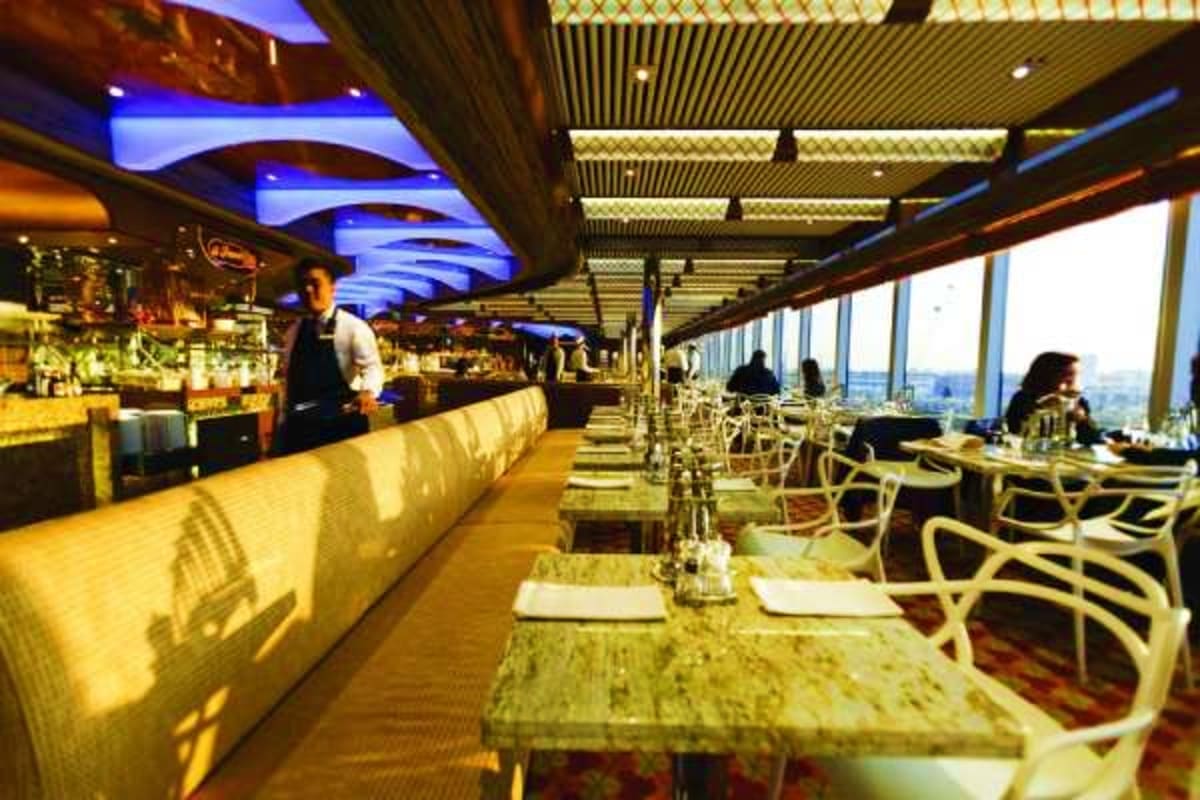 Modern cruise ship dining room with floor-to-ceiling windows overlooking ocean and coastal city, lit bar counter, and elegant