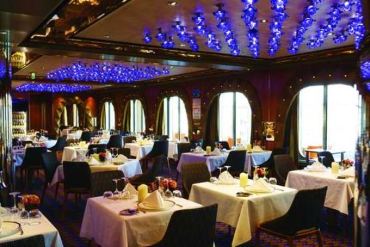 Elegant ship dining room with white tablecloth tables, black chairs, large windows, and blue LED ceiling lights on Costa Diad